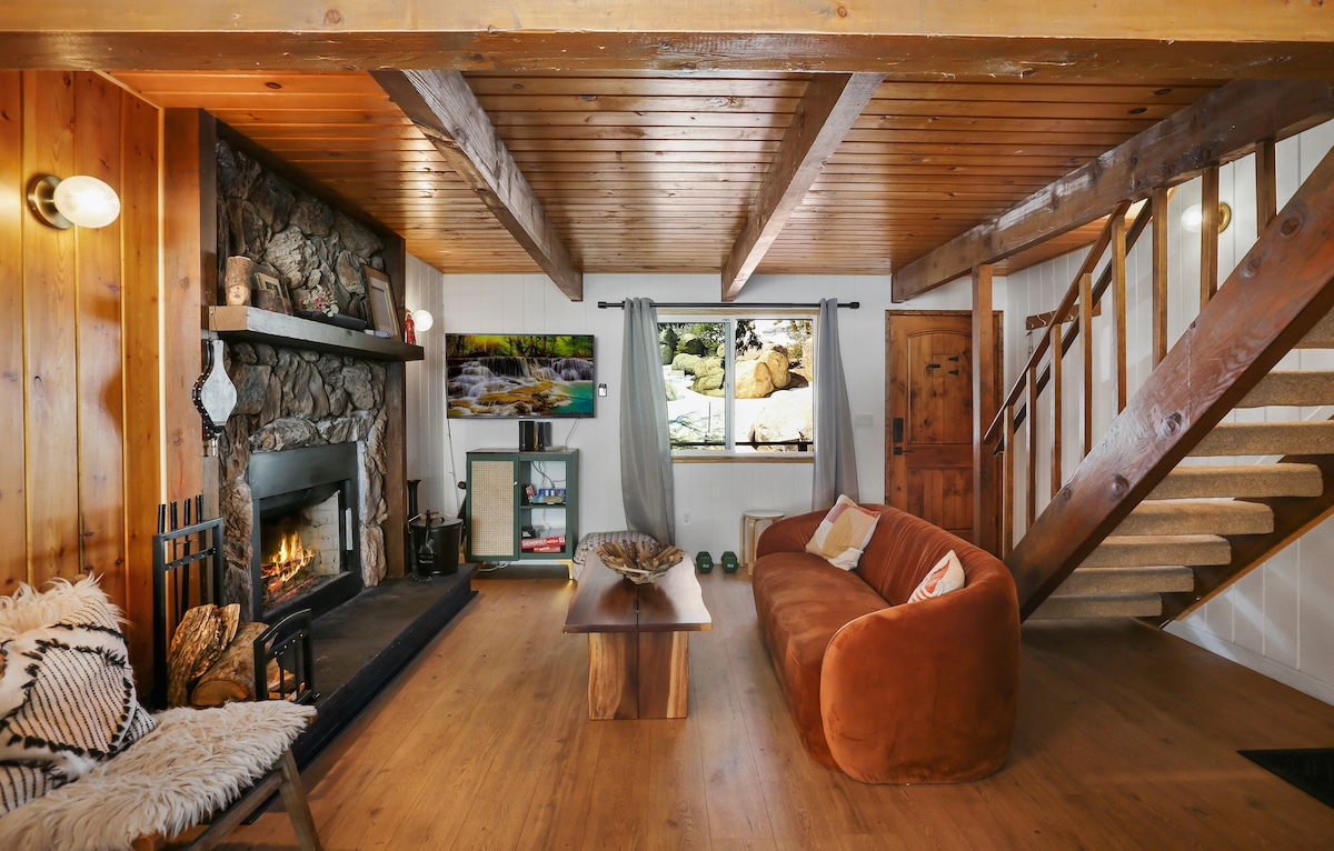 A warm living area features a stone fireplace surrounded by wooden walls and a ceiling with exposed beams. A cozy brown sofa sits adjacent to a wooden coffee table, while a television screen displays a serene nature scene. Wooden stairs lead to the upper level.