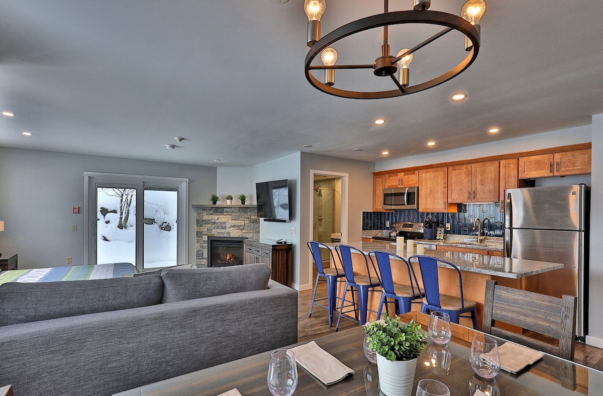 An open living area is depicted, featuring a cozy sofa facing a gas fireplace. A well-equipped kitchen is highlighted with stainless steel appliances and a spacious island with seating for six. Large glass doors provide access to a snowy outdoor view.