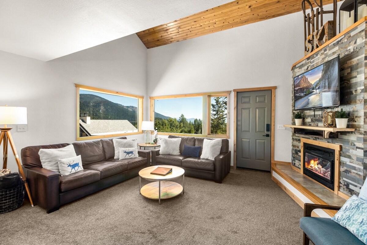 The spacious living room features high ceilings with wooden beams. A cozy dark leather sofa is arranged around a circular coffee table. Large windows allow natural light and showcase views of the surrounding mountains. A stone fireplace and a wall-mounted television enhance the warm atmosphere.