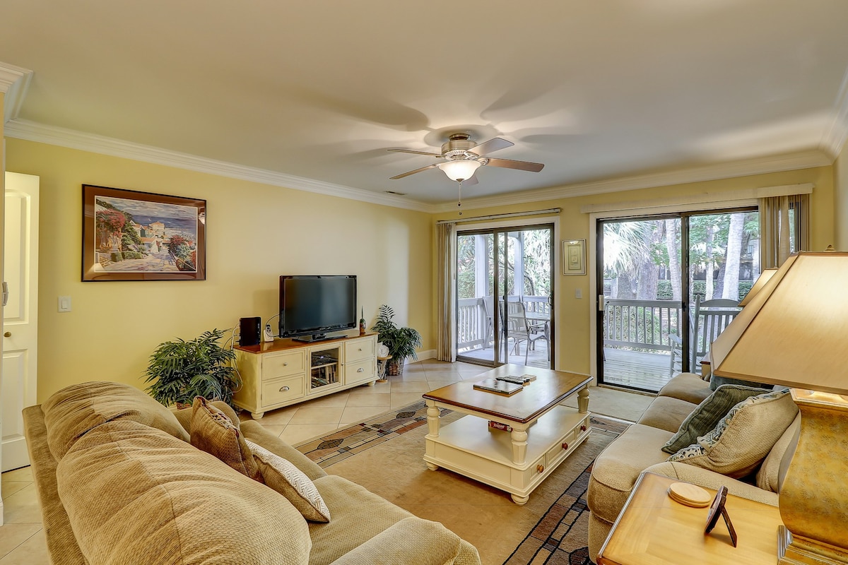 A spacious living area features light tile flooring and soft beige sofas arranged around a coffee table. A flatscreen TV is positioned on a light wooden media console. Large sliding glass doors provide access to the outdoor porch, allowing natural light to fill the room.