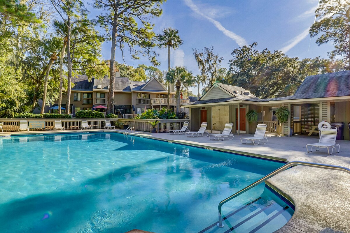 A clear swimming pool is surrounded by a spacious deck featuring several lounge chairs. Lush trees and greenery add to the tranquil atmosphere, with buildings visible in the background, offering easy access to the pool area.