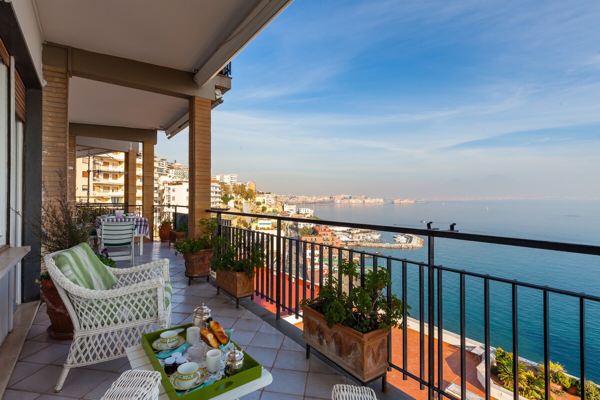 A terrace offers panoramic views of the Gulf of Naples, featuring potted plants and outdoor seating. A small table set for breakfast is visible, with a selection of pastries and beverages, while the water reflects a clear blue sky.