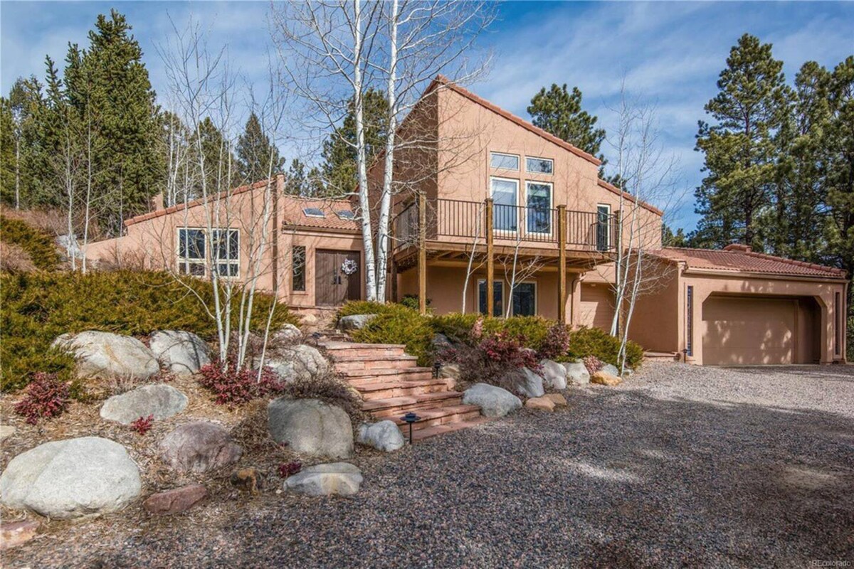 The exterior of a spacious home is depicted with a warm earth-toned facade, featuring large windows that invite natural light. Surrounding greenery includes bushes and tall trees, enhancing the setting's serenity. A pathway leads to the entrance, framed by smooth stones and gravel.