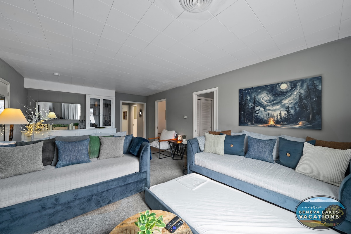 A bright living area is furnished with two spacious sofas adorned with a mix of blue and neutral cushions. A round coffee table is positioned at the center, while a large artwork with a starry night scene hangs above. Soft light from a nearby lamp illuminates the room.