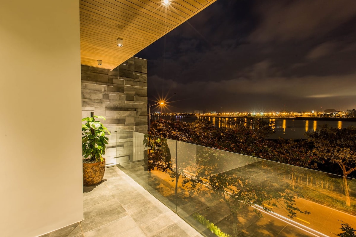 A terrace area is observed with sleek tile flooring and a glass railing overlooking a river under a night sky. Soft lighting highlights the space, complemented by a potted plant that adds a touch of greenery. The city lights are visible across the water.