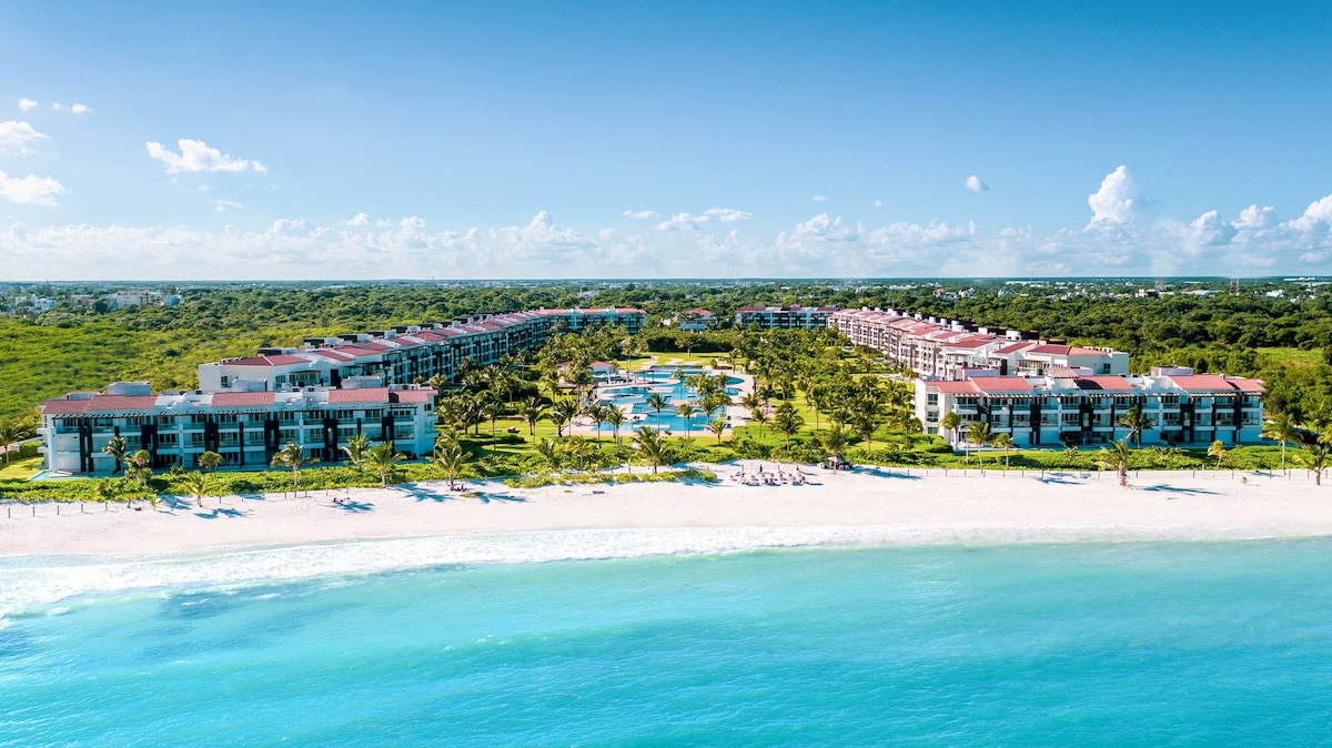Aerial view of the Mareazul Resort displays its lush green landscaping and well-maintained beach. The expansive infinity pool sits centrally, surrounded by palm trees and accommodations. The coastline features sandy beach areas and turquoise waters, reflecting clear blue skies dotted with light clouds.