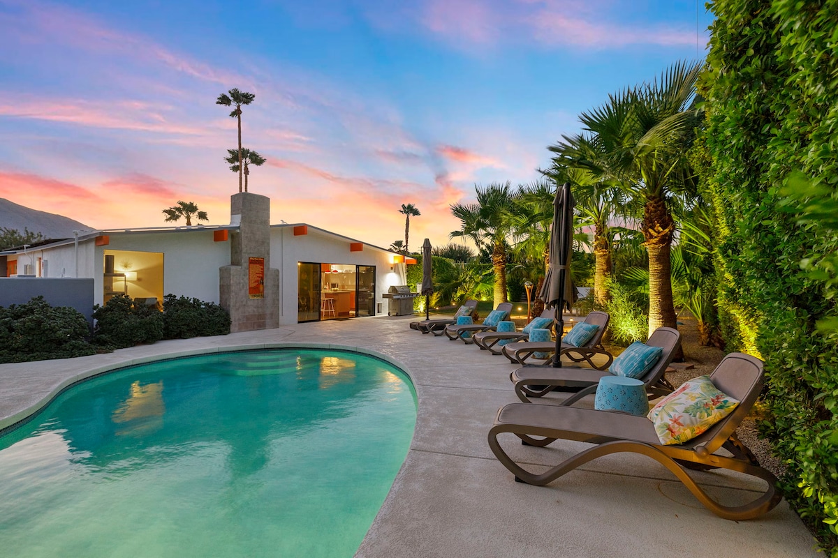 A spacious patio features a kidney-bean shaped pool surrounded by lounge chairs, each with decorative cushions. Lush palm trees and landscaping provide privacy, while a modern home can be seen in the background, under a colorful sky at dusk.