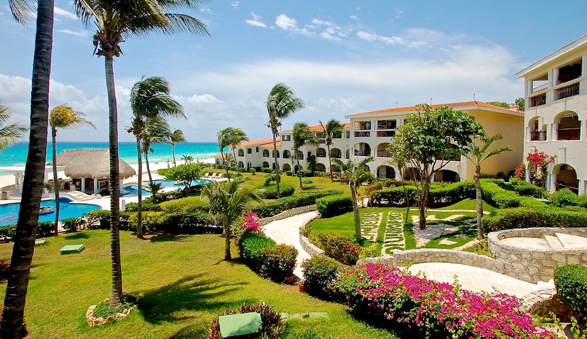 The image captures the vibrant outdoor grounds of the Xaman Ha complex, showcasing manicured gardens interspersed with colorful flowers. Palm trees sway gently in the breeze, while the ocean and white sandy beach are visible in the background. The pool area is also featured, with lounge chairs positioned around the inviting water.