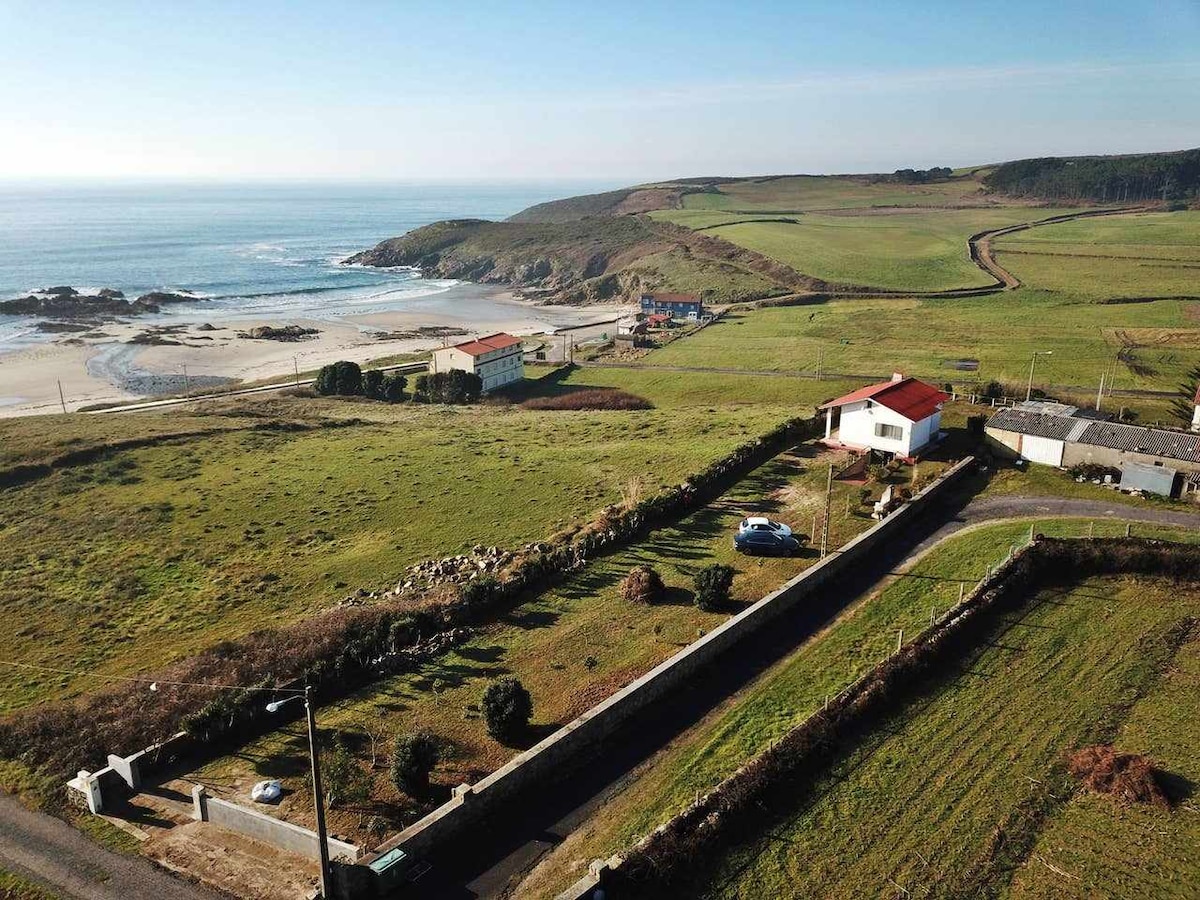 An aerial view captures the expansive green landscape surrounding the coastal area, showcasing fields and a distant beach. A white building with a red roof is visible, along with a few neighboring structures. The ocean glistens under clear skies, enhancing the serenity of the location.
