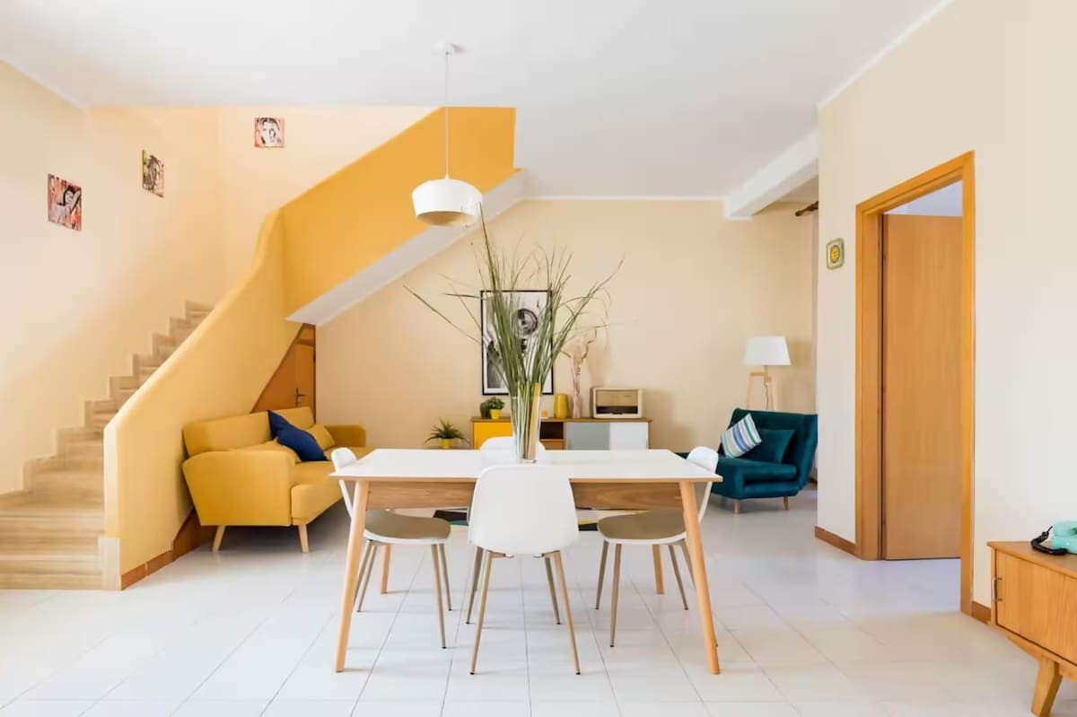 An inviting dining area features a white table surrounded by six chairs in various colors. A yellow accent wall adds brightness, and a staircase with wooden steps leads to the upper level. A yellow sofa and a teal armchair provide comfortable seating nearby.