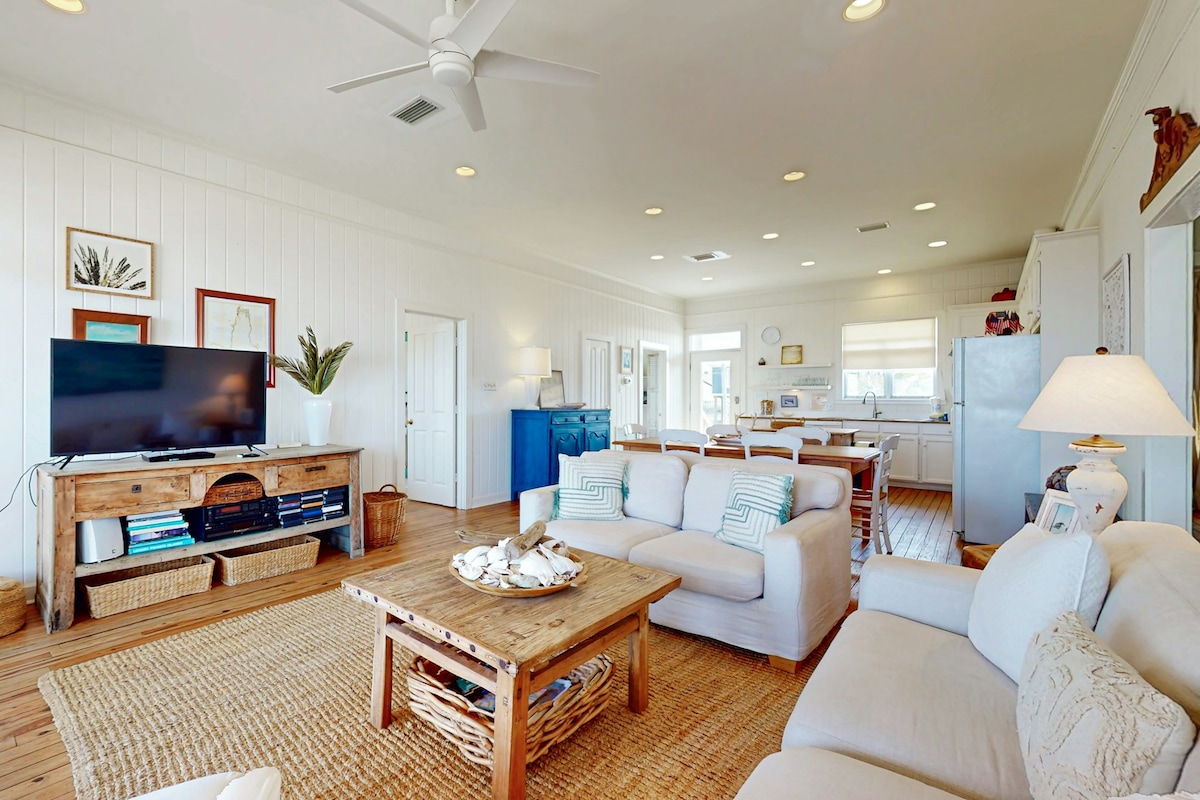 An open living area features light-colored furnishings, including a spacious sectional sofa and a wooden coffee table adorned with seashells. A television set is mounted on a rustic console, while a dining space with wooden chairs and a blue sideboard can be seen in the background.