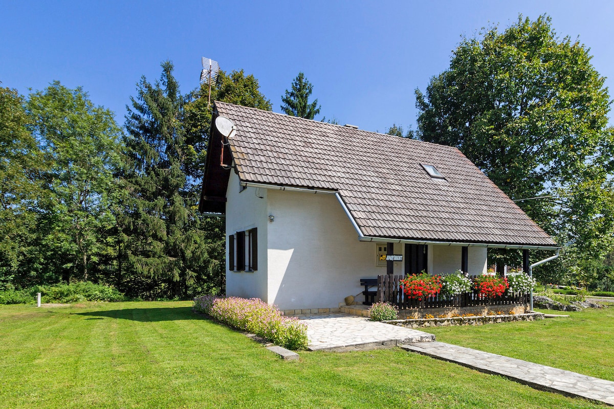 The holiday home is set amidst a green landscape, featuring a sloped roof and white exterior. A flower garden with vibrant blooms is positioned at the front, while large trees surround the property, contributing to a peaceful atmosphere.