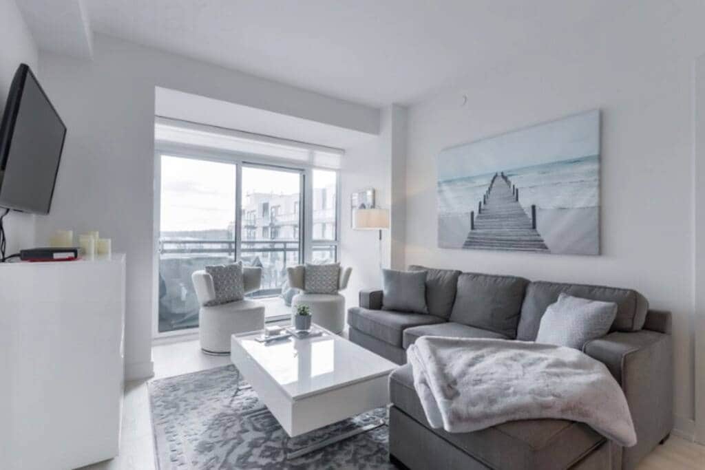 The living area features a gray sectional sofa and a modern coffee table placed on a patterned rug. Two light-colored armchairs add seating options. A large artwork depicting a serene pathway leads to water decorates the wall, while a balcony with glass doors offers ample natural light.