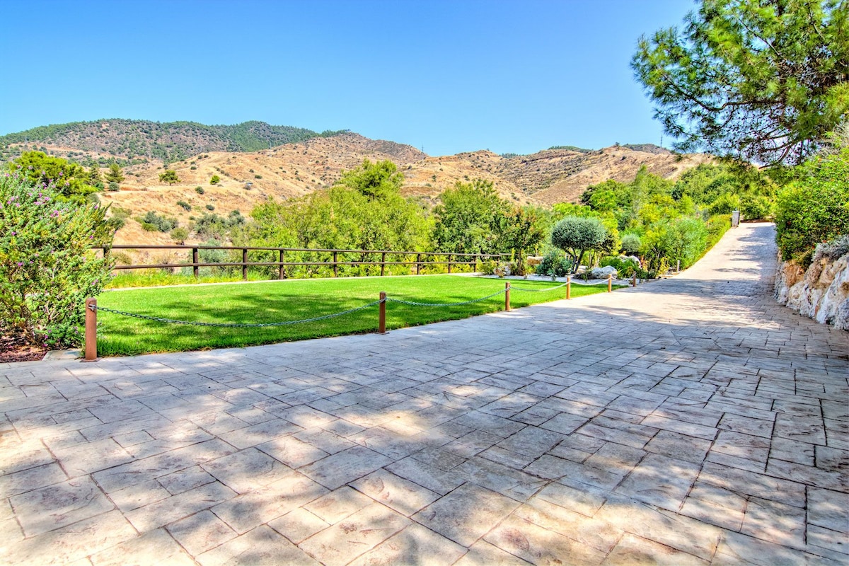 A spacious driveway leads through landscaped gardens, bordered by lush, green lawns and trees. The panoramic views of mountains in the background complement the serene landscape. Sunlight bathes the area, enhancing the inviting surroundings.