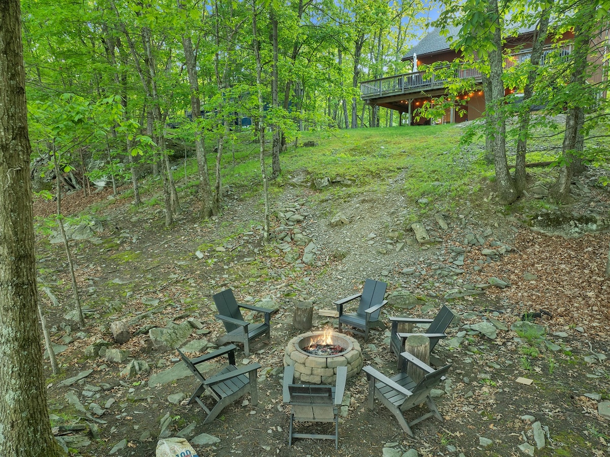 An outdoor setting showcases a firepit surrounded by multiple black Adirondack chairs. The area is bordered by trees, creating a natural, peaceful atmosphere. In the background, a cabin is visible with a deck overlooking the scene.