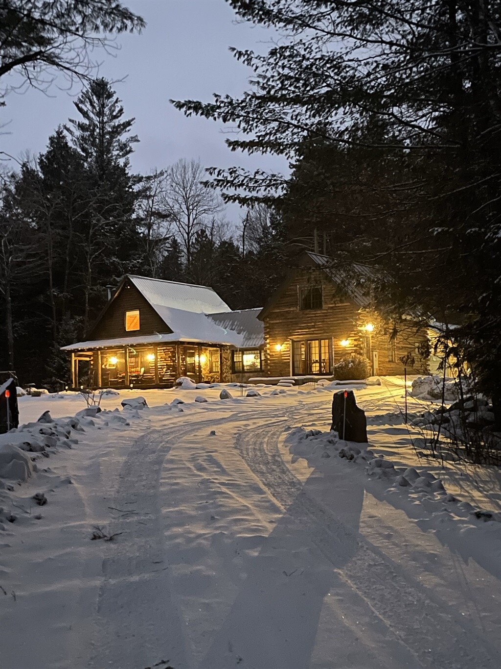 A snowy pathway leads to a charming cabin illuminated by warm lights, highlighting its rustic log structure. The landscape is adorned with a fresh layer of snow, surrounded by tall trees, creating a serene setting as dusk approaches.