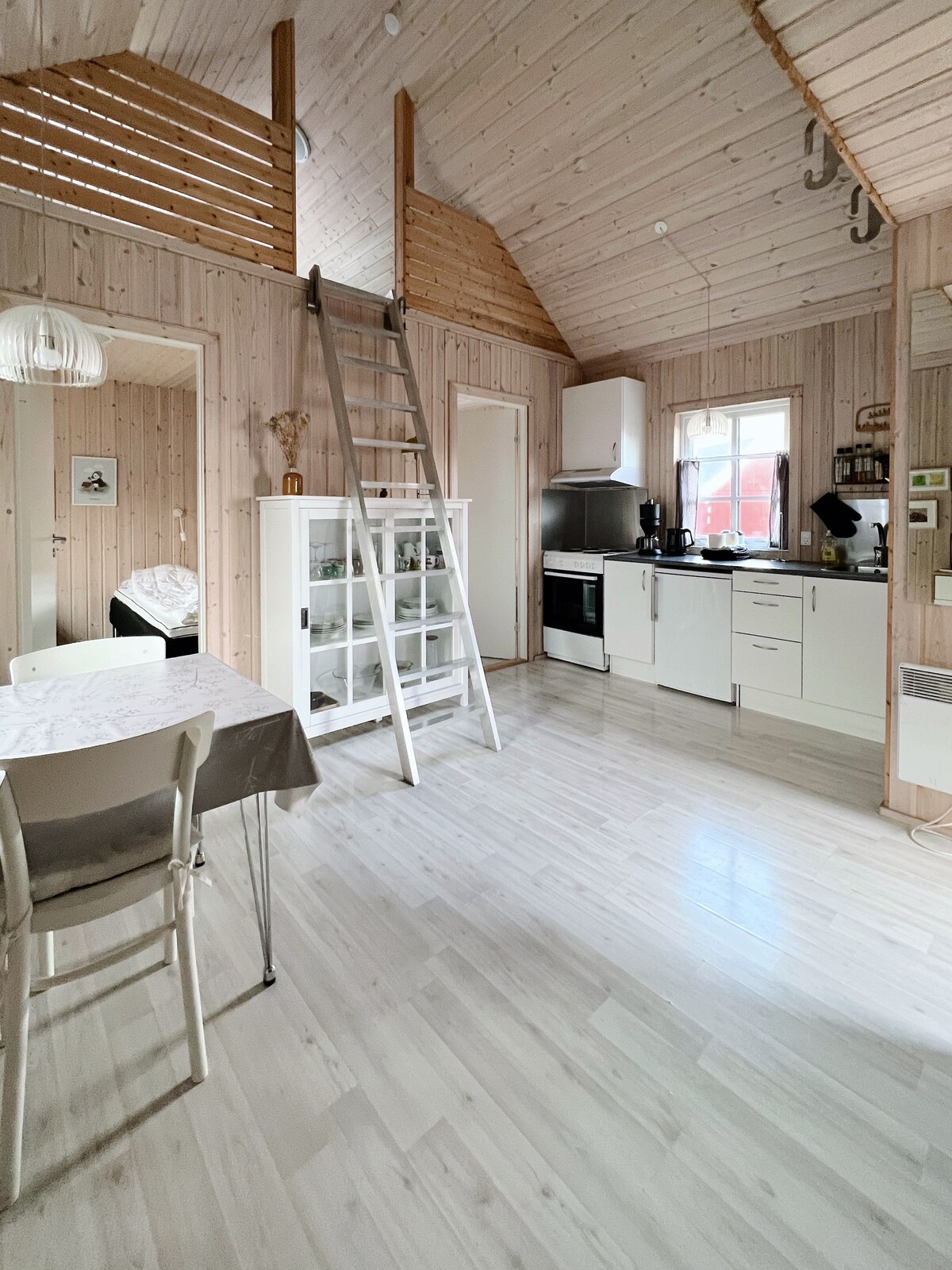 An open-plan living space features wooden walls and vaulted ceilings, creating a light and airy feel. A dining area with a table and chairs is positioned to the left, while a kitchenette with modern appliances is in the background. A ladder leads to a sleeping loft above.