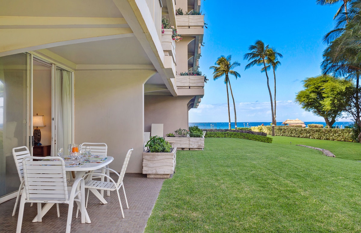 An expansive wrap-around balcony features a dining table and chairs, set against a backdrop of lush green lawn and swaying palm trees. Partial views of the ocean are visible in the distance, complementing the serene outdoor space.