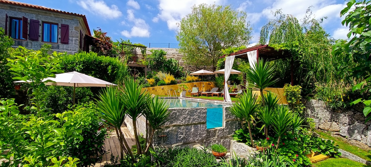 A granite pool surrounded by lush greenery and vibrant flowers is featured. Sun loungers are positioned beside the water, while shaded areas with umbrellas provide comfort. The tranquil garden is accentuated by tall plants and a large tree, creating a serene atmosphere.