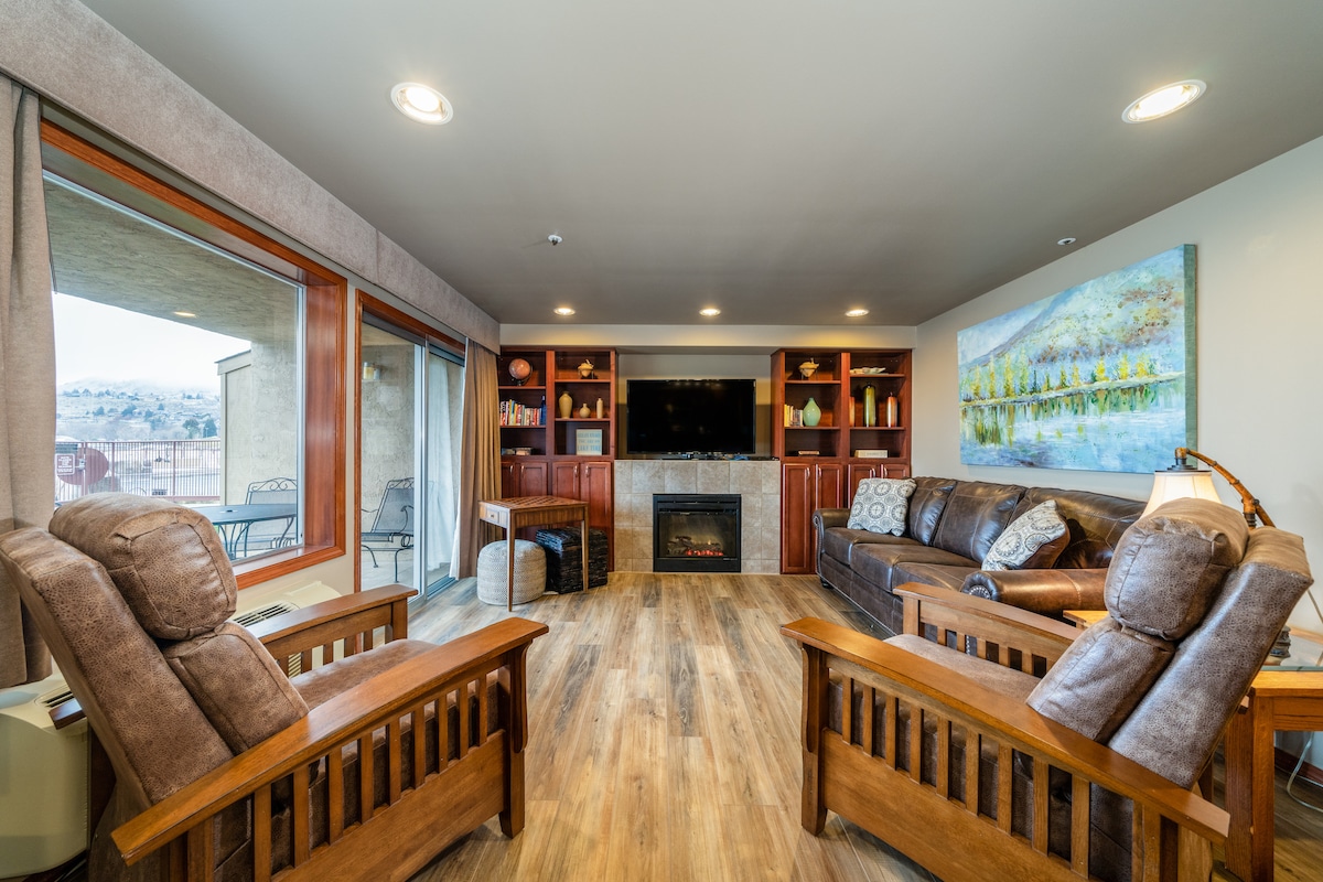 A well-appointed living area features a comfortable seating arrangement with two wooden recliners and a spacious leather sofa. Natural light enters through large windows, illuminating a modern fireplace and built-in shelves adorned with decorative items. A serene landscape painting enhances the calming atmosphere.
