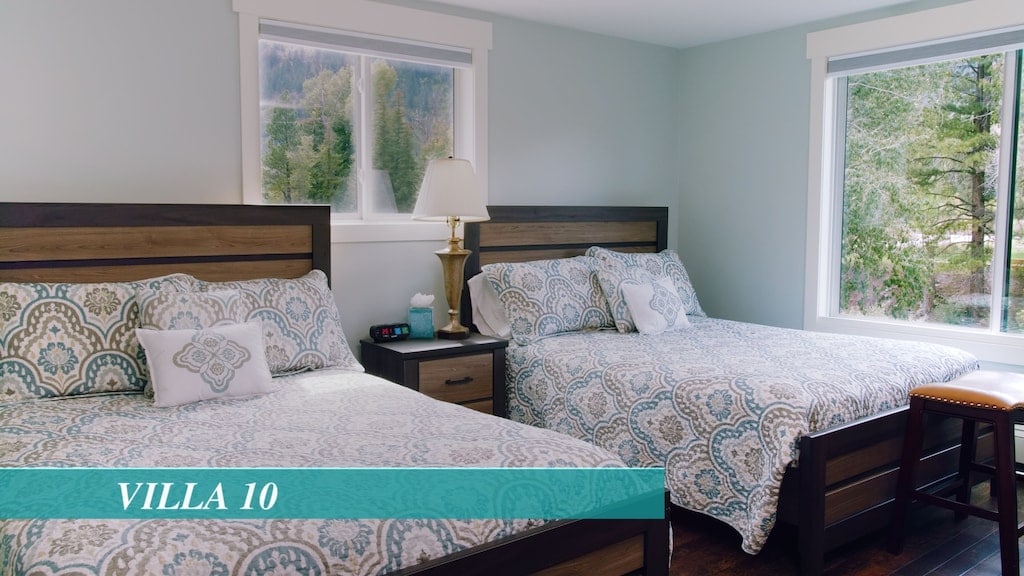 A well-appointed bedroom features two spacious beds, each adorned with patterned covers. Nightstands with lamps accompany the beds, providing functional space. Large windows allow natural light to fill the room, with views of the surrounding greenery visible through the glass.