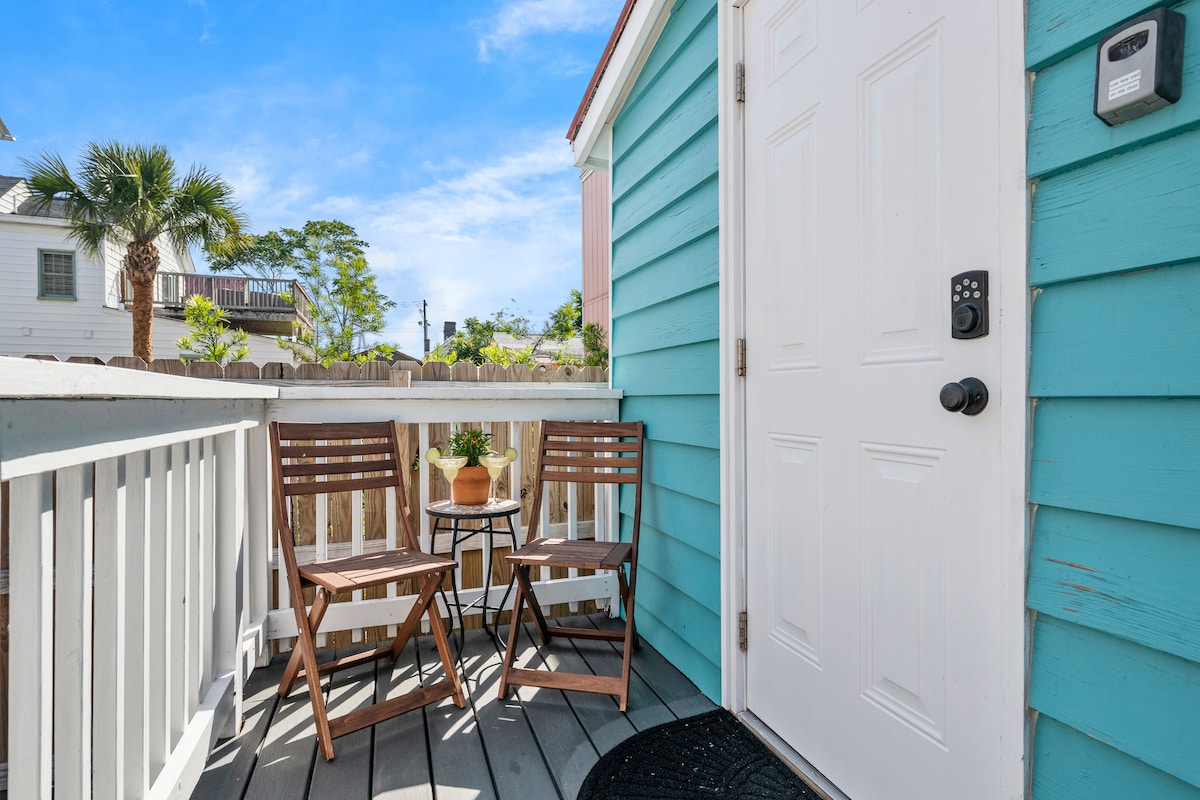 A bright entrance features a light blue exterior with a white door and modern keypad lock. Two wooden chairs and a small table create a cozy seating area, complemented by a potted plant. Sunlight brightens the space, enhancing the welcoming atmosphere.