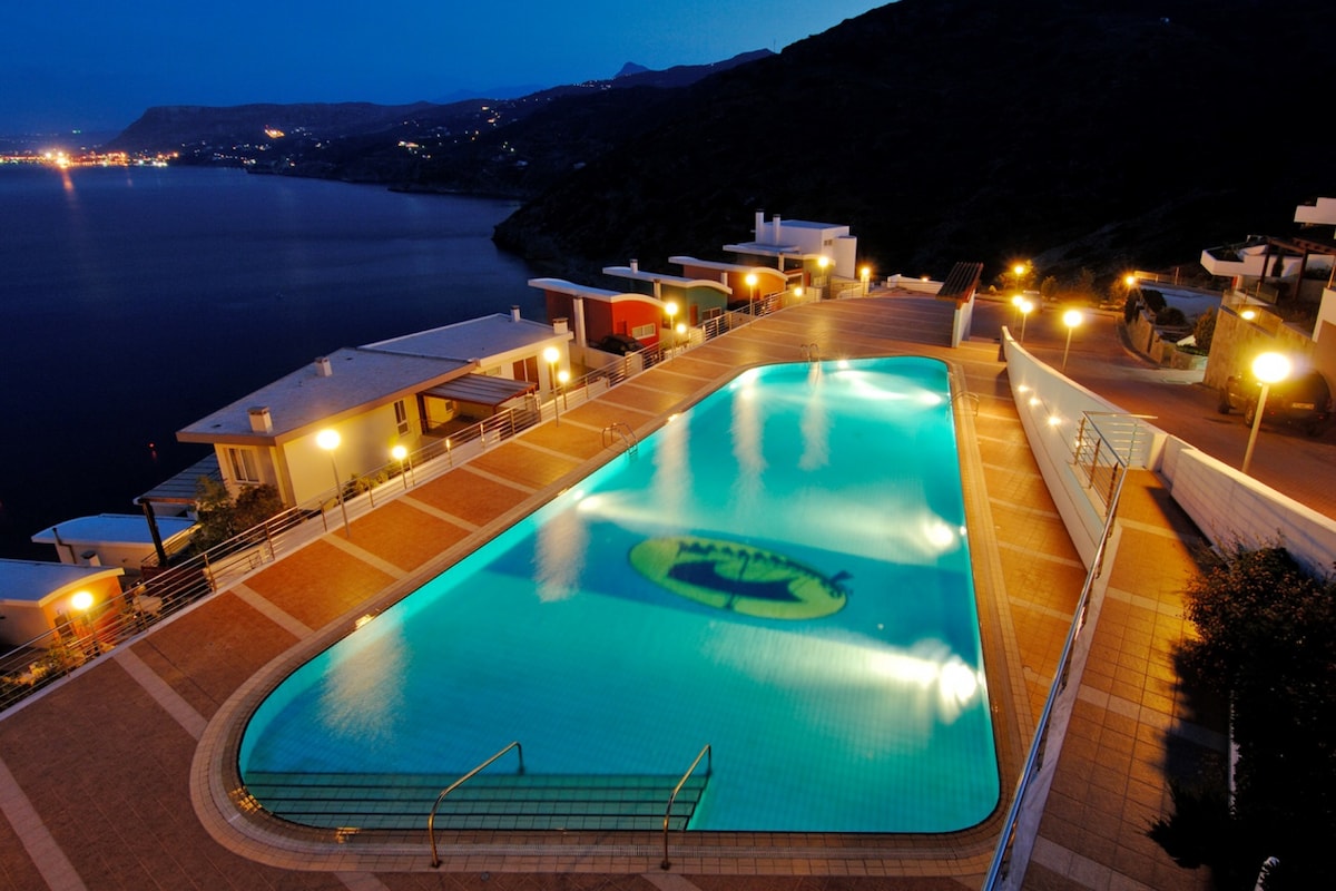 A spacious communal pool is illuminated at night, reflecting calming turquoise hues. Surrounding the pool are well-lit walkways and landscaped areas, with a panoramic view of the coastline and distant lights from neighboring homes visible in the background.