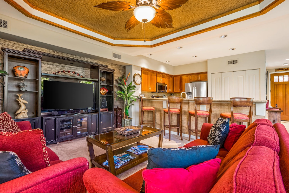 A spacious living area features a comfortable red sectional sofa and a glass coffee table. A large flat-screen TV is mounted on a dark wooden entertainment unit. The open-concept kitchen is visible in the background, equipped with wooden cabinetry and bar seating.