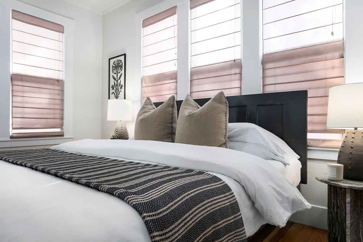 A studio sleeping area is presented with a spacious bed featuring a white duvet and decorative pillows. Natural light filters through sheer blinds on three windows, illuminating the warm wooden floor. Two bedside lamps with textured bases are positioned on either side of the bed.