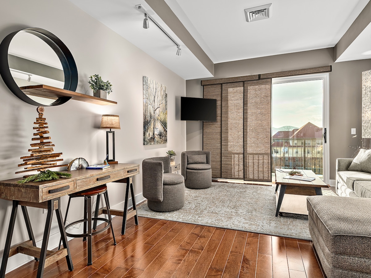 A modern living area features a cozy seating arrangement with two accent chairs and a spacious sofa. A wooden console table displays decorative items. Natural light enters through large sliding glass doors, leading to a balcony that offers views of the outdoors.