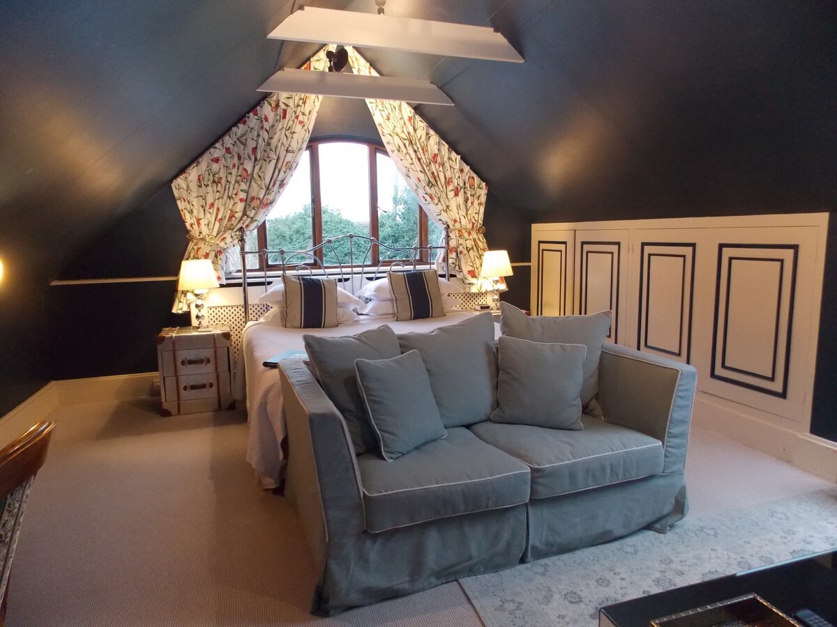 The loft-style room features a spacious bed adorned with soft linens, complemented by a cozy sofa in a calming shade. Large windows with floral drapes offer natural light and views of the surrounding greenery, enhancing the room's serene atmosphere.