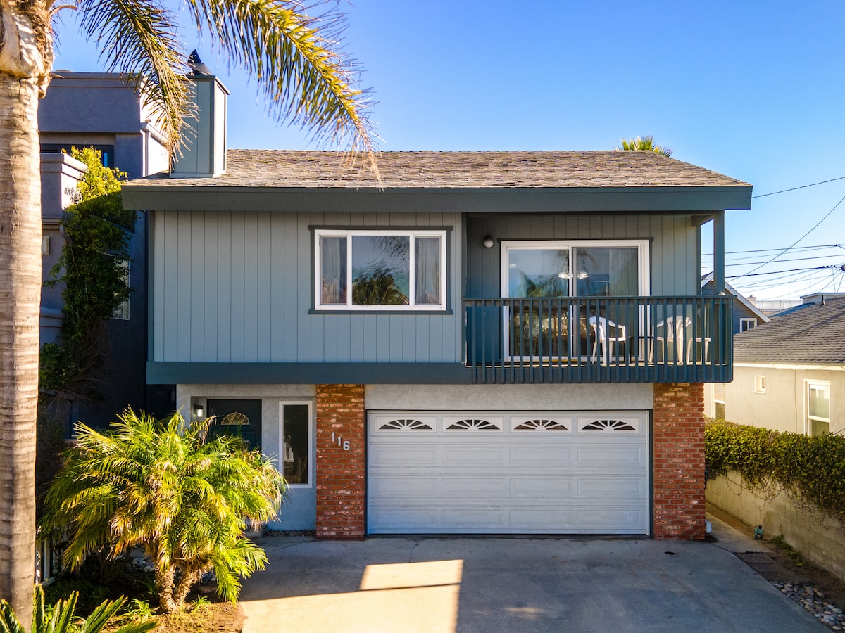 A two-level beach home is presented, featuring a light blue exterior and a spacious driveway. Palm trees frame the entrance, while a balcony is visible on the upper level, providing outdoor seating. The garage door beneath displays decorative arches, complementing the vintage charm of the home.