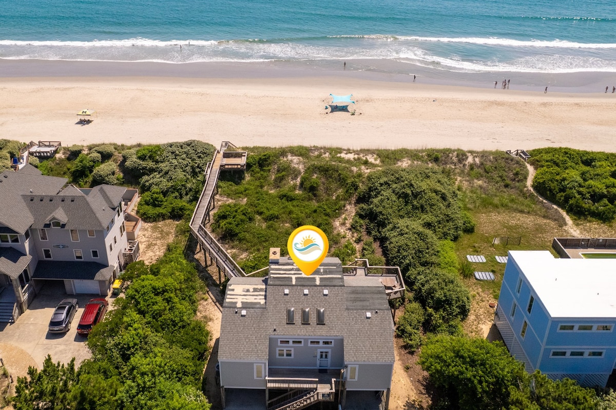 Aerial view features an oceanfront cottage surrounded by natural dunes and lush greenery. The private walkway leads directly to the beach, with soft sand and gentle waves visible in the distance. Nearby homes are set within a tranquil coastal landscape.