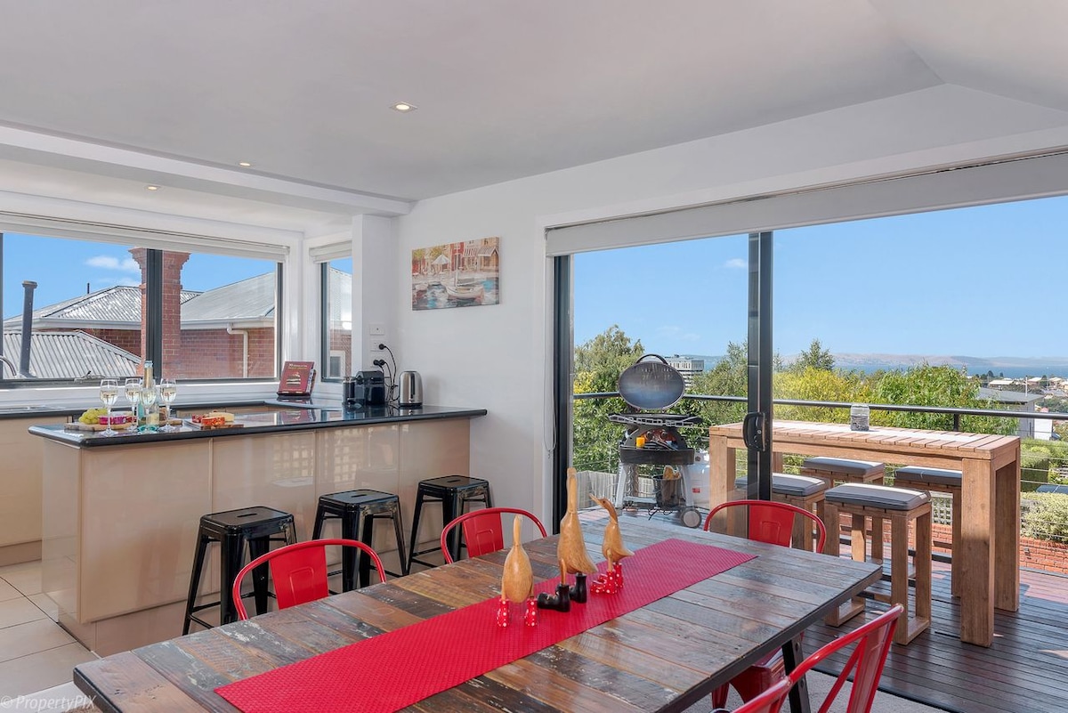A well-lit dining area features a large wooden table with red accents and black stools. The space offers expansive windows revealing water views and access to an outdoor deck equipped with a BBQ. A range of kitchen appliances is neatly arranged on the counter.
