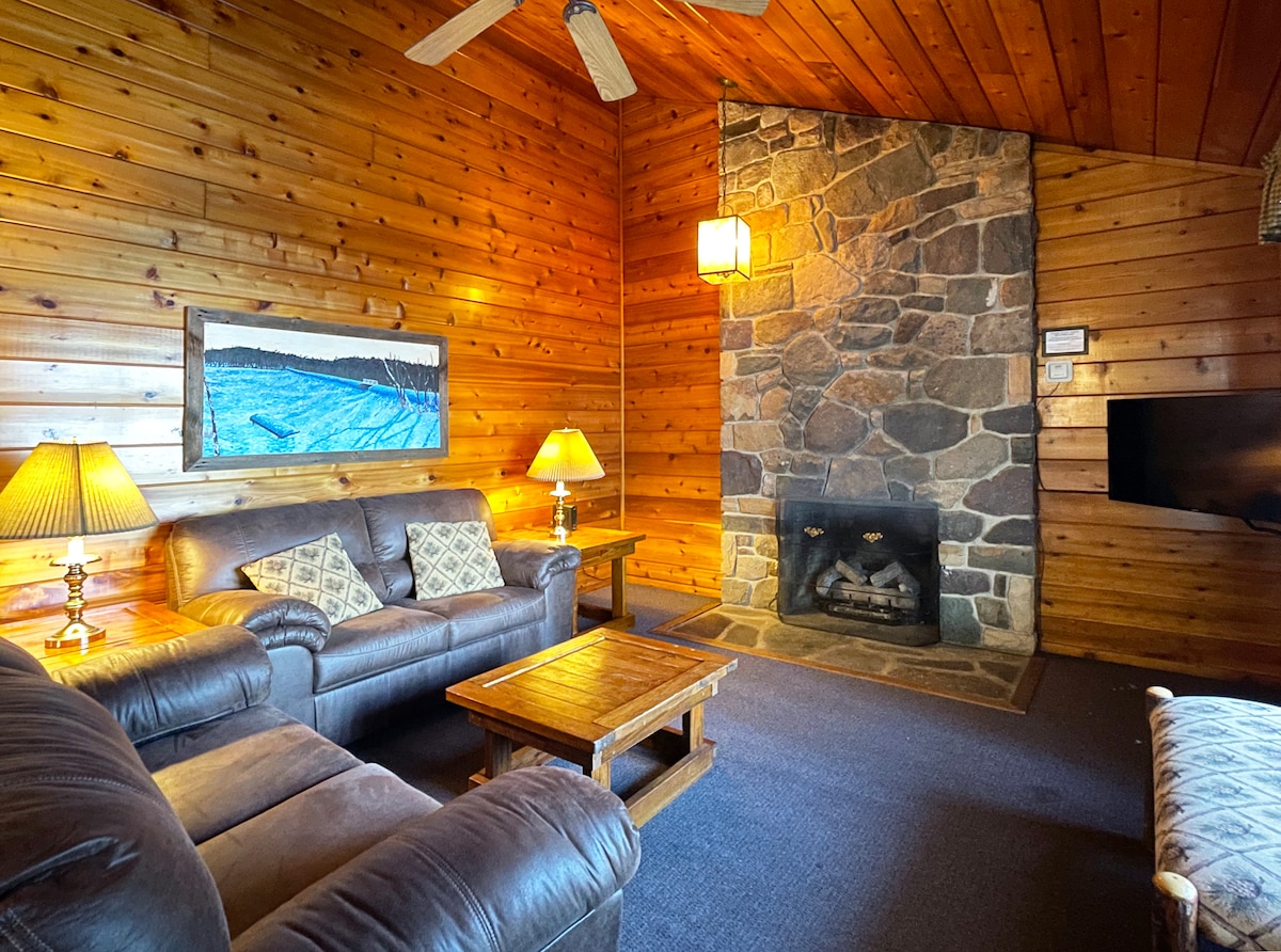 A cozy living area features warm wooden walls and a grand stone fireplace, complemented by two soft couches. A wooden coffee table sits centrally, surrounded by lamps that provide a soft glow, while a wall-mounted television enhances the comfort of the space.