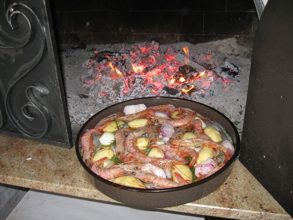 A metal tray filled with shrimp and vegetables rests on a stone countertop, positioned in front of glowing embers. The red and orange hues of the fire reflect on the tray, creating a warm ambiance. The background shows blackened bricks, enhancing the cooking environment.