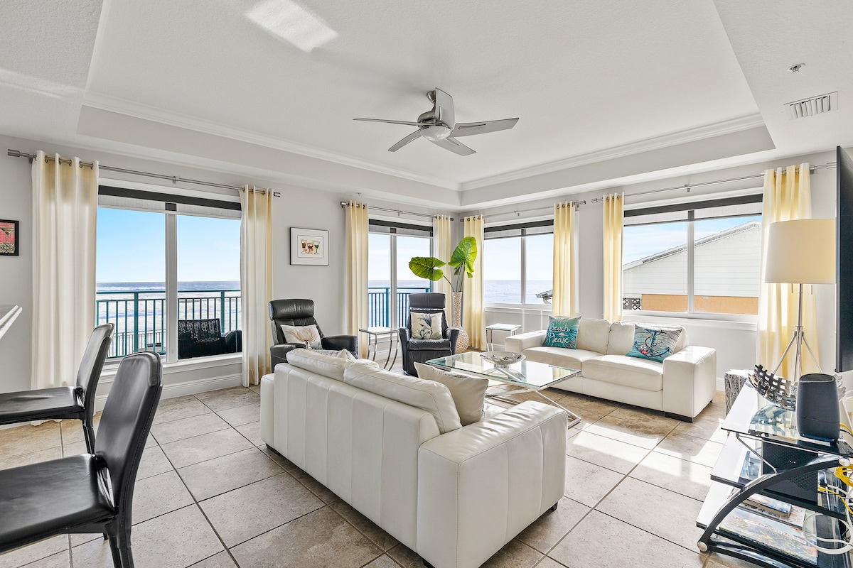 The living area features large windows offering sweeping ocean views. A combination of white sofas, a sleek glass coffee table, and a ceiling fan enhances the space. Light-colored curtains frame the windows, and potted greenery adds a touch of vibrancy to the room.