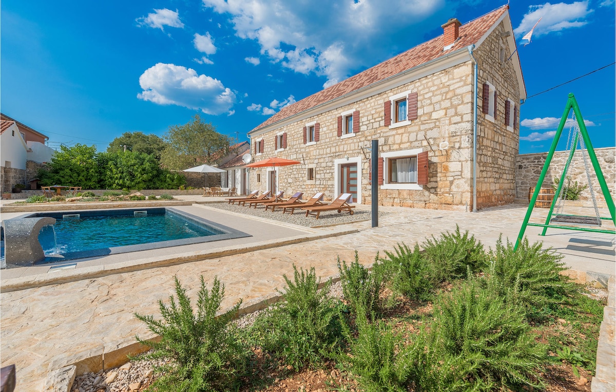 An exterior view of a charming stone house is presented, featuring a large outdoor pool surrounded by sun loungers. Lush greenery and native plants adorn the area, with a swing set positioned to the right. Clear blue skies enhance the inviting atmosphere.