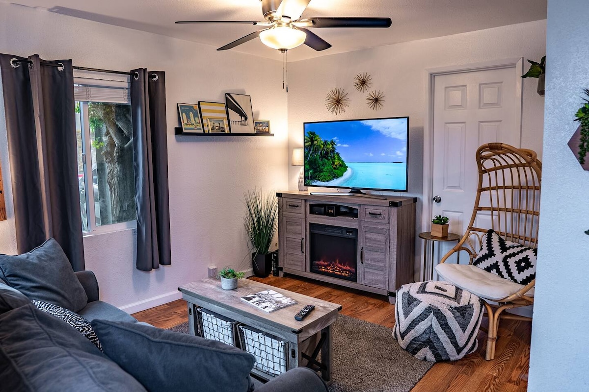The living room features a comfortable seating area with a large sectional couch facing a flat-screen Smart Roku TV. A cozy electric fireplace adds warmth, while natural light enters through a nearby window adorned with light curtains. Stylish decor elements include a patterned pouf and modern wall art.