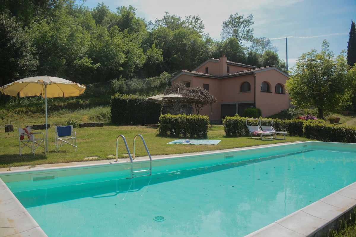 A private swimming pool with turquoise water is framed by a green lawn. Sun loungers and umbrellas are arranged nearby, providing space for relaxation. The villa is visible in the background, surrounded by trees and greenery, creating a serene atmosphere.