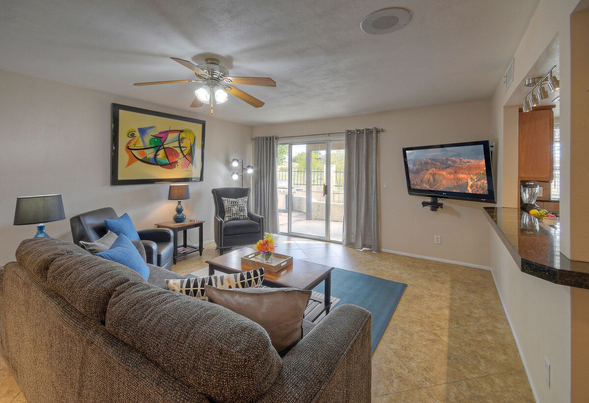The spacious living area features a comfortable sectional sofa and two armchairs, arranged around a coffee table. A large Smart TV is mounted on the wall, with natural light flowing through sliding glass doors that open to the patio. Decorative art and lamps enhance the ambiance.