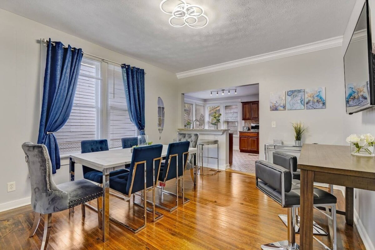 A dining area features a sleek table surrounded by six stylish chairs, complemented by a large window draped with blue curtains. A wall-mounted television and a small bar table are visible, providing a modern and functional space for meals and gatherings.