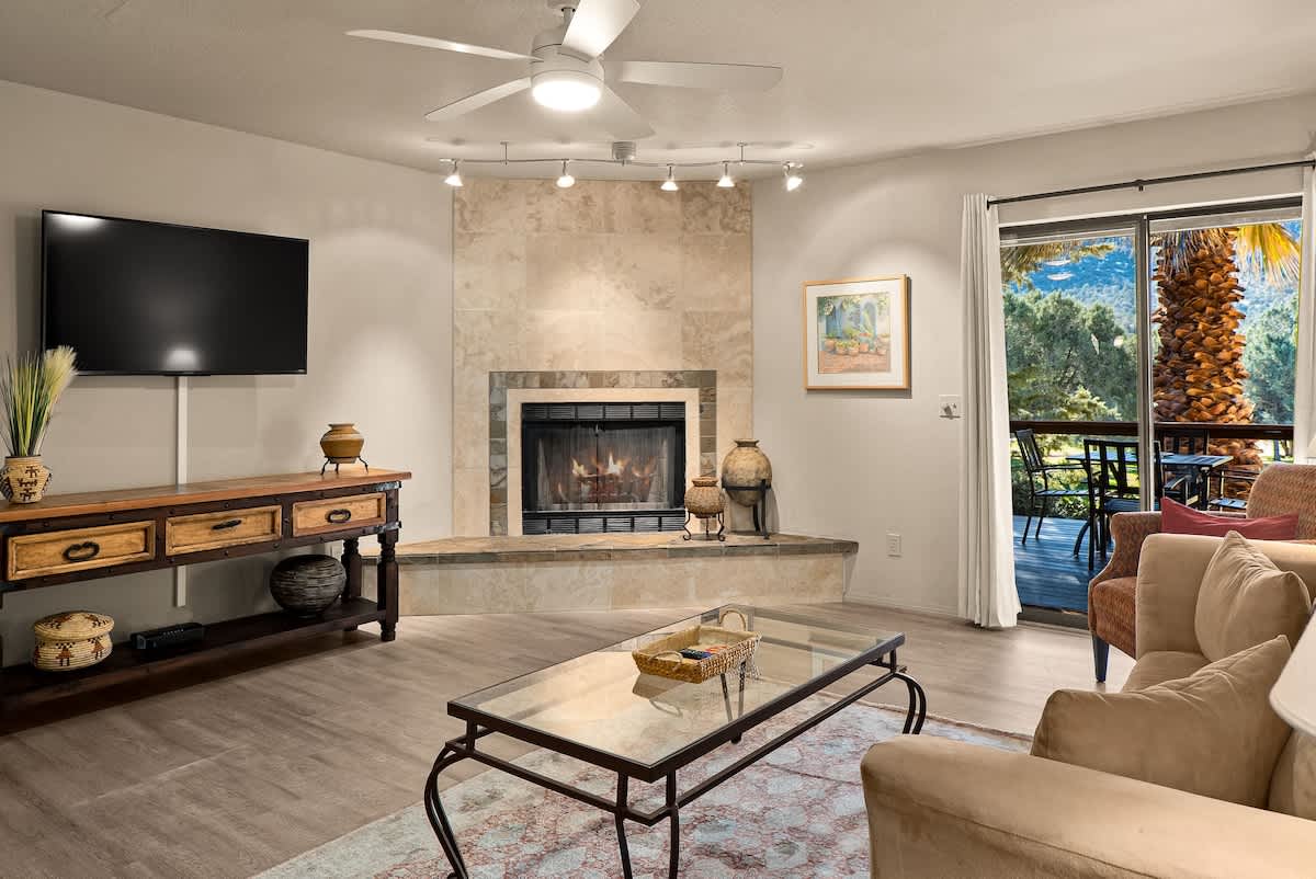A cozy living space is highlighted by a mounted television and a fireplace with a stone surround. A comfortable couch is positioned in view of a glass coffee table. Natural light flows through large sliding doors leading to the private patio, where outdoor seating is visible.