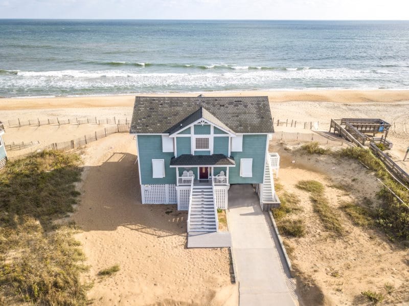 A beachfront vacation home is seen from above, showcasing its teal exterior and welcoming staircase leading to the entrance. The sandy beach extends along the ocean, with gentle waves lapping at the shore, and a pathway visible leading to the beach access.