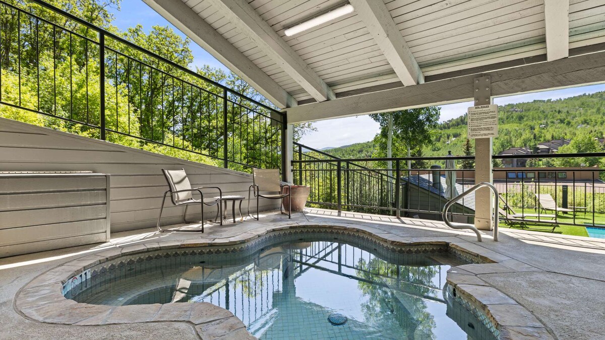 A hot tub is situated under an open-air roof, surrounded by a spacious deck. Two lounge chairs are positioned near the edge, while lush greenery is visible in the background, enhancing the sense of relaxation and connection to nature.