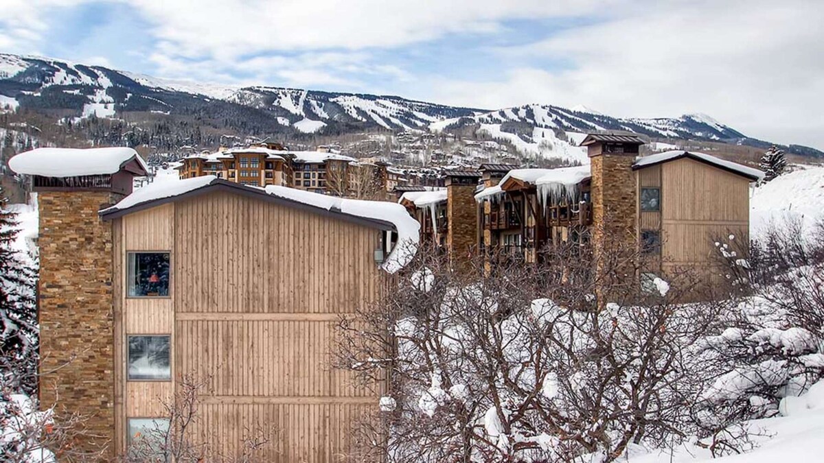 The exterior of the condo is shown, featuring a wood and stone facade against a backdrop of snow-covered mountains. The surrounding landscape includes trees blanketed in snow, creating a serene winter atmosphere. The ski slopes are visible in the distance under a partly cloudy sky.