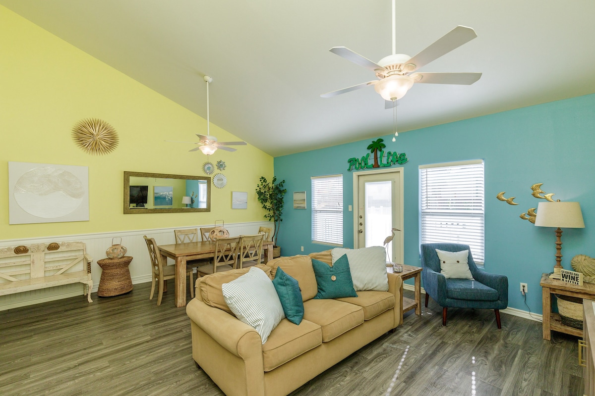 The bright and airy living area features a beige sofa adorned with blue and green accent pillows. A dining table for six is positioned under a mirrored wall with light fixtures. Brightly colored accent walls in yellow and blue enhance the open-concept space, illuminated by natural light.