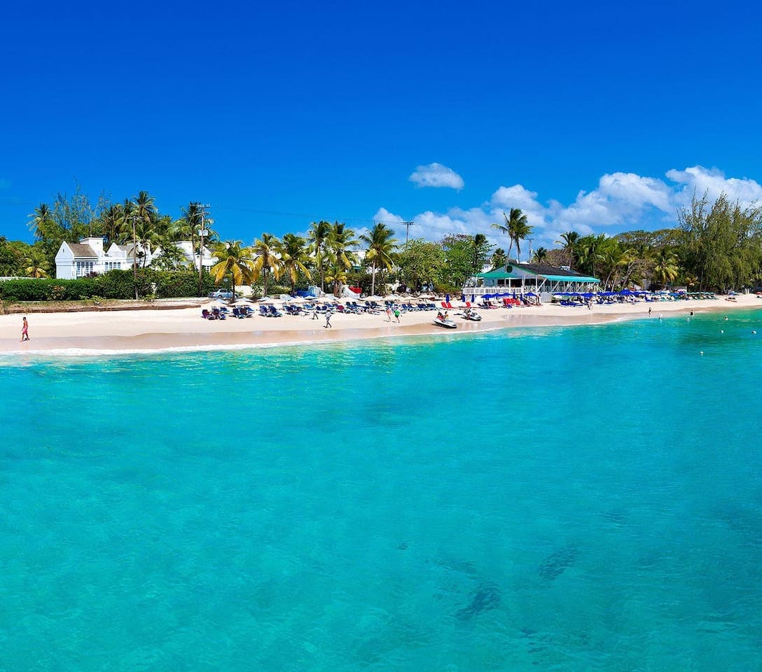 A sandy beach stretches along the shoreline, with gentle waves lapping at the water’s edge. Sun loungers and umbrellas are arranged under palm trees, while a clear turquoise sea reflects the bright blue sky. Scenic tropical vegetation lines the background.