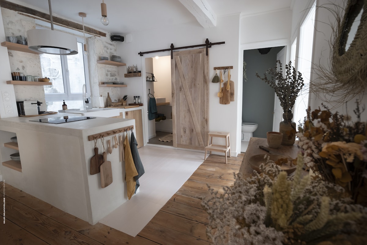 The kitchen features a modern design with an open layout and warm wooden flooring. A wooden island with a sink stands at the center, while a sliding barn door leads to the bathroom area. Decorative plants and kitchen utensils are displayed on the wall, enhancing the inviting ambiance.