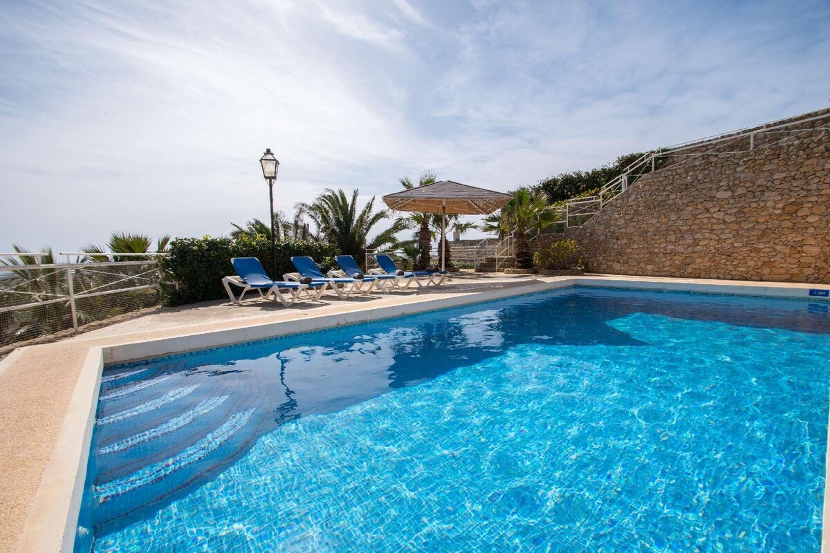 A spacious raised swimming pool area is highlighted, featuring a clear blue pool surrounded by sun loungers. Palm trees and a shaded gazebo are visible in the background, providing a relaxed outdoor atmosphere. The area is enclosed with a stone wall, enhancing privacy.